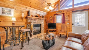 A cozy cabin interior with a stone fireplace, a television, a leather sofa, and a dining area with a glass table and metal chairs. - Dog Friendly Cabin, Sleeps 2・Hot Tub・Near Park (Pigeon Forge)