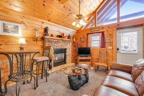 A cozy cabin interior with a stone fireplace, a television, a leather sofa, and a dining area with a glass table and metal chairs. - Dog Friendly Cabin, Sleeps 2・Hot Tub・Near Park (Pigeon Forge)
