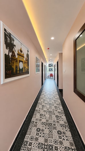 A corridor with patterned flooring, framed artwork on the walls, and a red chair in the distance. - Hotel Singh Palace (Varanasi)