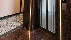 Un ascensor moderno con puerta de cristal, rodeado de paredes de mármol oscuro y una lámpara decorativa.