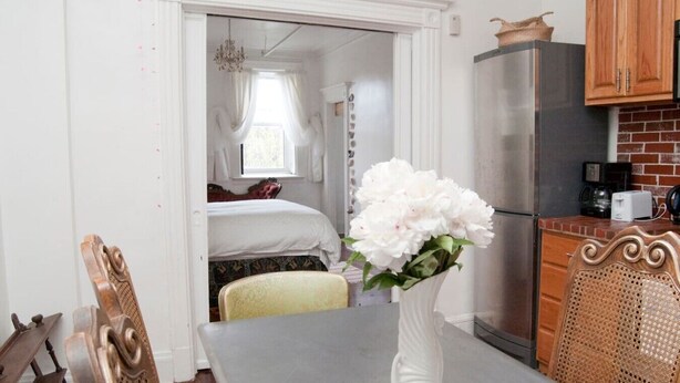A room with a dining table, a white vase with flowers, a bed with a window, and a kitchen area with a refrigerator and wooden cabinets.