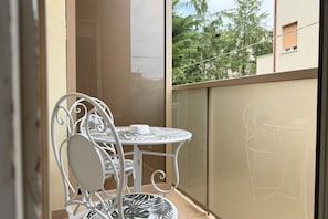 Ein Balkon mit einem weißen Metalltisch und Stühlen, einem kleinen weißen Tisch sowie Blick auf Bäume und Gebäude.