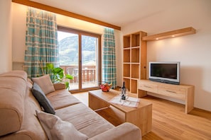 Una sala de estar con un sofá, una mesa de centro, un televisor sobre un soporte de madera y un balcón con cortinas.