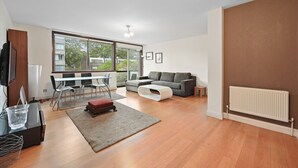 Ein modernes Wohnzimmer mit einer Couch, einem Couchtisch, einem Fernseher, einem Esstisch mit Stühlen und einem großen Fenster.