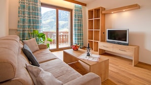 Ein Wohnzimmer mit einer Couch, einem Couchtisch, einem Fernseher auf einem Holzboden und einem Balkon mit Vorhängen.