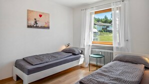 A single bed with a gray mattress, a small table, a radiator, and a window with a view of greenery and a building. - Dog-friendly, modern holiday apartments with terrace, 2 balconies and Wi-Fi (Bräunlingen)