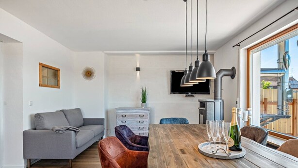 A modern living room with a grey sofa, a wooden dining table with wine glasses and a bottle, a TV mounted on the wall, and a window with a view of the outside. - Dog-friendly, modern holiday apartments with terrace, 2 balconies and Wi-Fi (Bräunlingen)