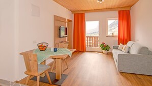 Een kamer met een houten plafond, een eettafel met stoelen, een grijze bank, een televisie en een raam met gordijnen.