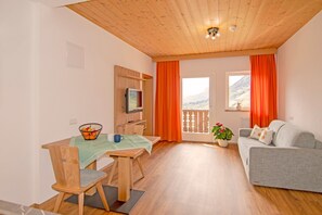 Ein Zimmer mit Holzboden, einem Esstisch mit Stühlen, einem grauen Sofa, einem Fernseher und einem Fenster mit Vorhängen.