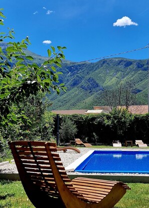 Una piscina con tumbonas de madera, cielo despejado y montañas al fondo.