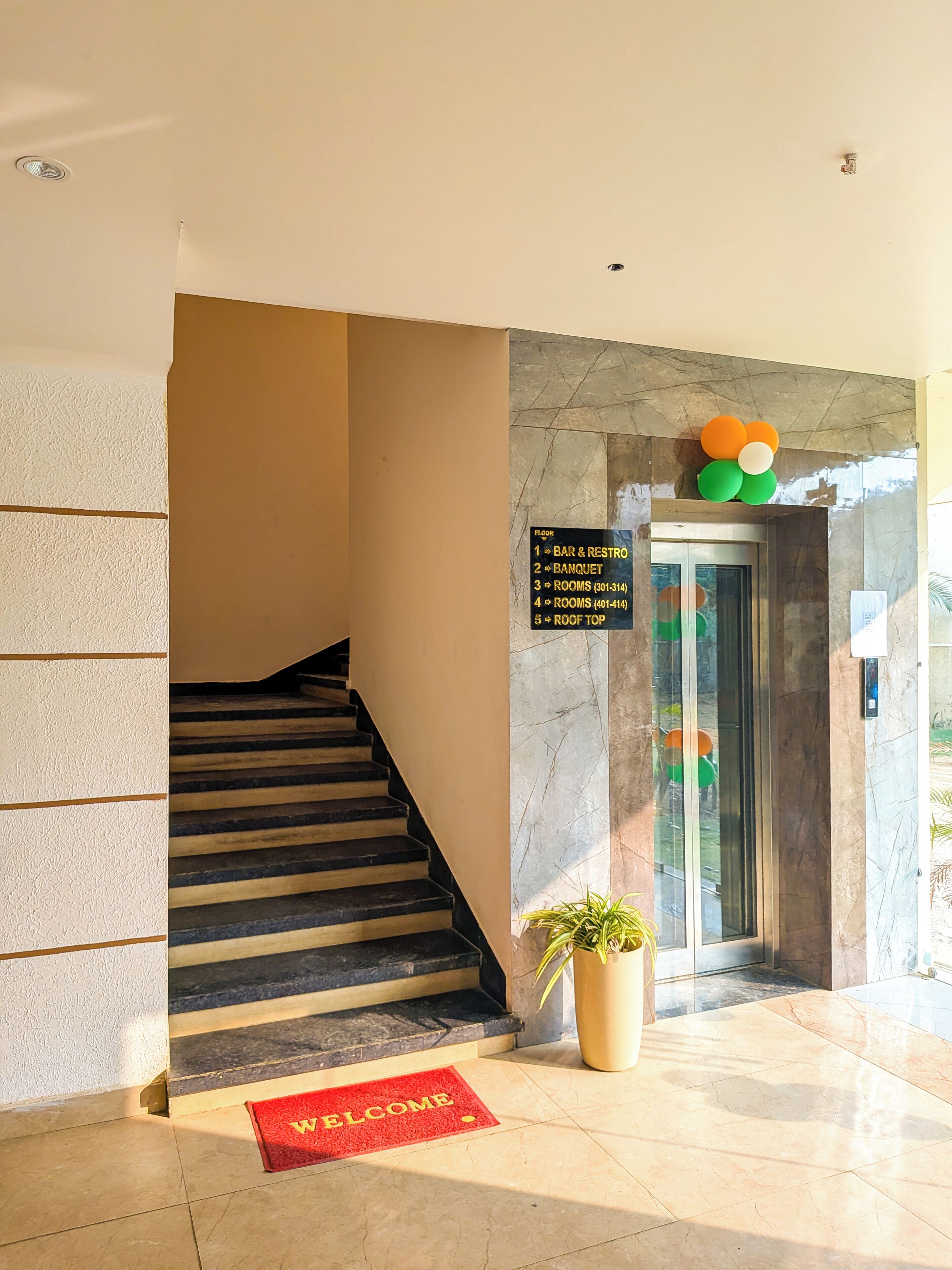 Um hall de hotel com uma escadaria, um tapete de boas-vindas vermelho, piso de mármore, um elevador de vidro e uma placa indicando números de quartos e andares.