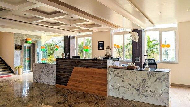 Un vestíbulo de hotel con mostrador de recepción, piso de mármol, una escalera y grandes ventanales con vistas a zonas verdes.