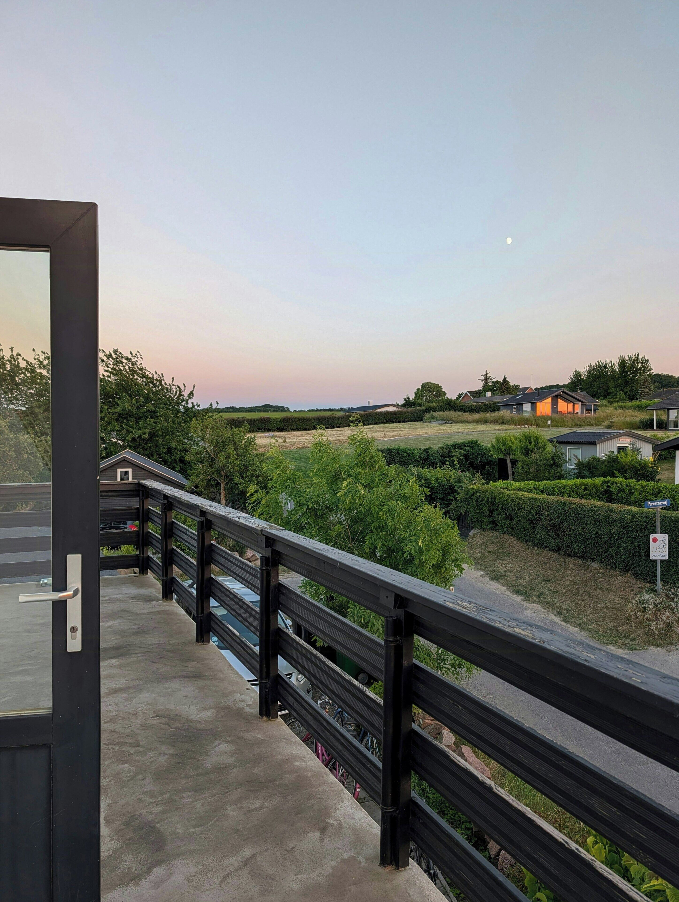 A view from a balcony with a wooden railing, overlooking a residential area with houses and greenery.