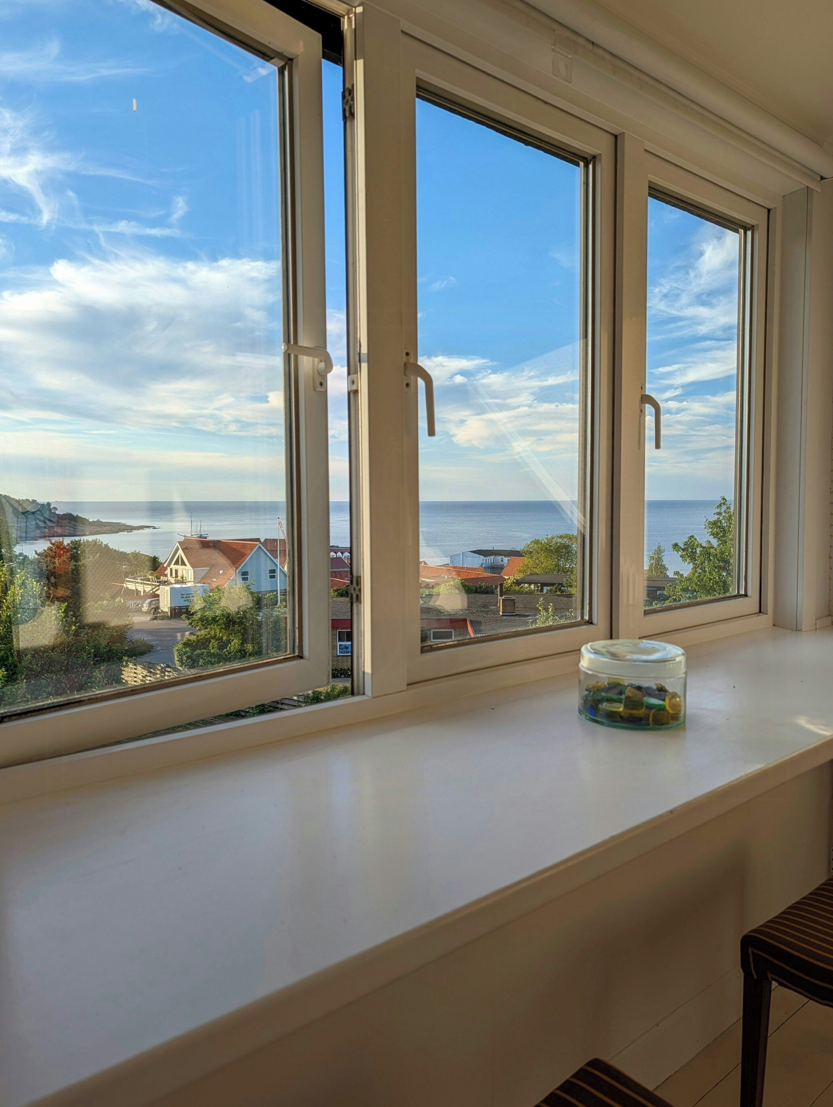 A room with three large windows overlooking a coastal area, a small table with a container on it, and a chair.