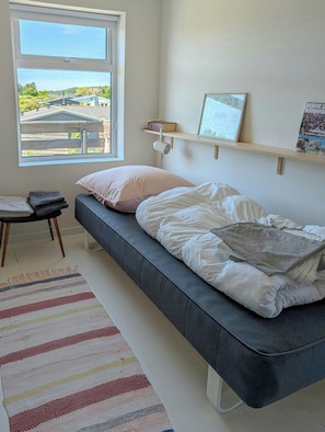 A bedroom with a bed, a window with a view, a shelf with books and a framed picture, and a small table with towels. - Semi-detached house with sea view in Sandvig (Allinge-Sandvig)