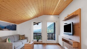Una sala de estar moderna con techo de madera, un sofá gris, un televisor montado en una pared de madera y un balcón con vista a la montaña.
