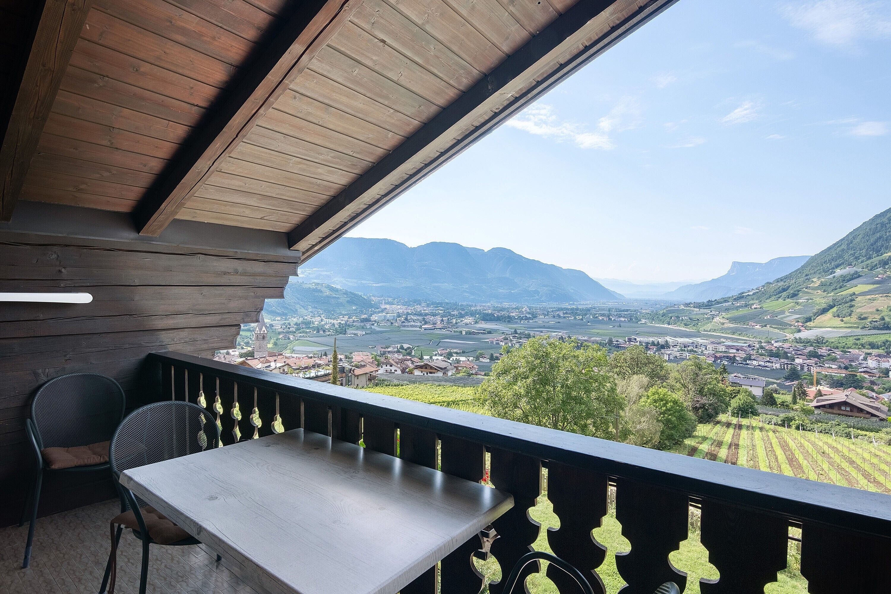 Un balcone con tavolo e sedie in legno, affacciato su una valle con montagne e una città.