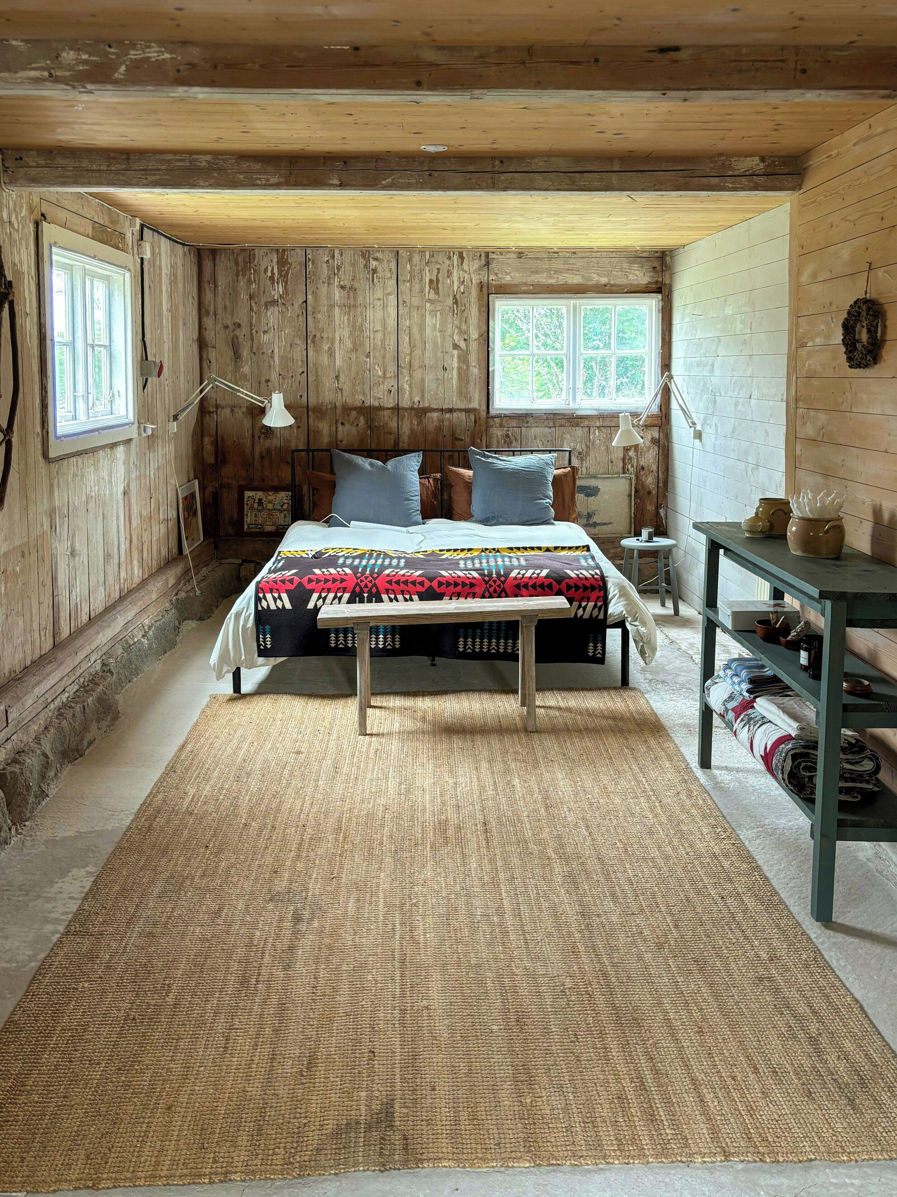 A rustic bedroom with a wooden ceiling, a bed with patterned bedding, two bedside lamps, a small table with decorative items, and a shelf with folded textiles.