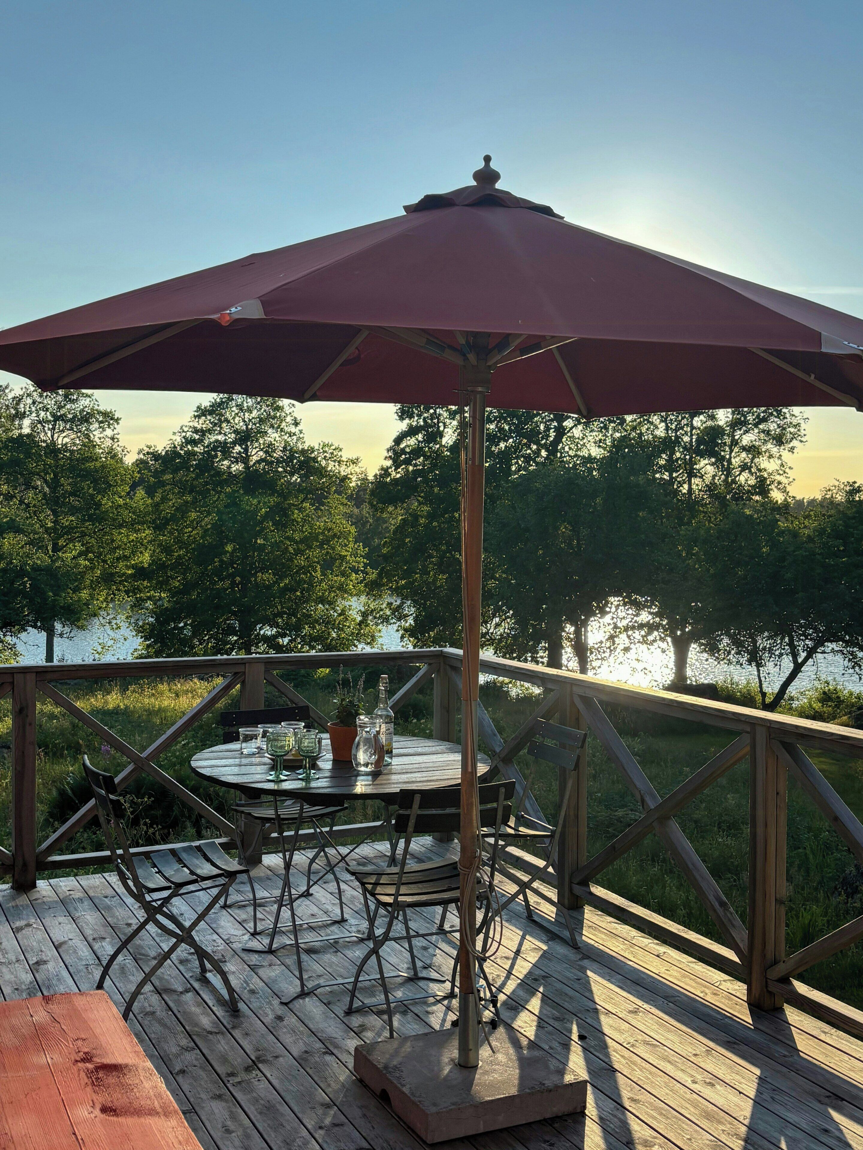 A wooden deck with a round table and chairs, an umbrella, and a view of trees and water.