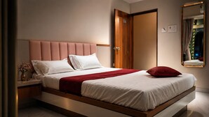 A modern bedroom with a large bed, a wooden headboard, a nightstand with a vase, and a mirror on the wall. - Hotel krushna palace lodge  (Wadwani)