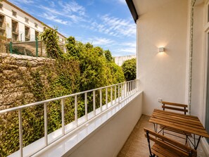 Ein Balkon mit weißer Reling, Holztischen und einer Steinmauer mit Pflanzenbewuchs.