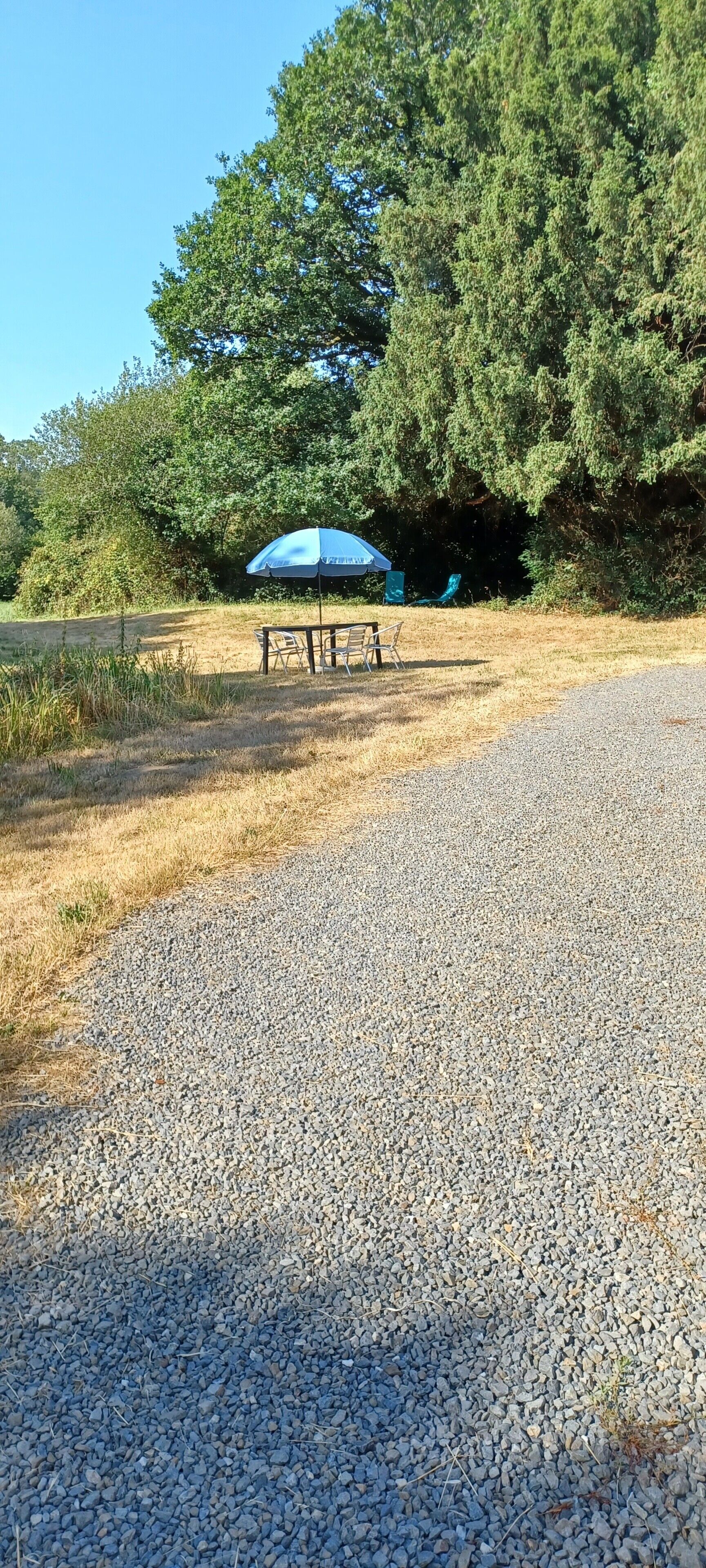 Un chemin de gravier mène à un espace aménagé avec des sièges, un parasol bleu et des chaises.