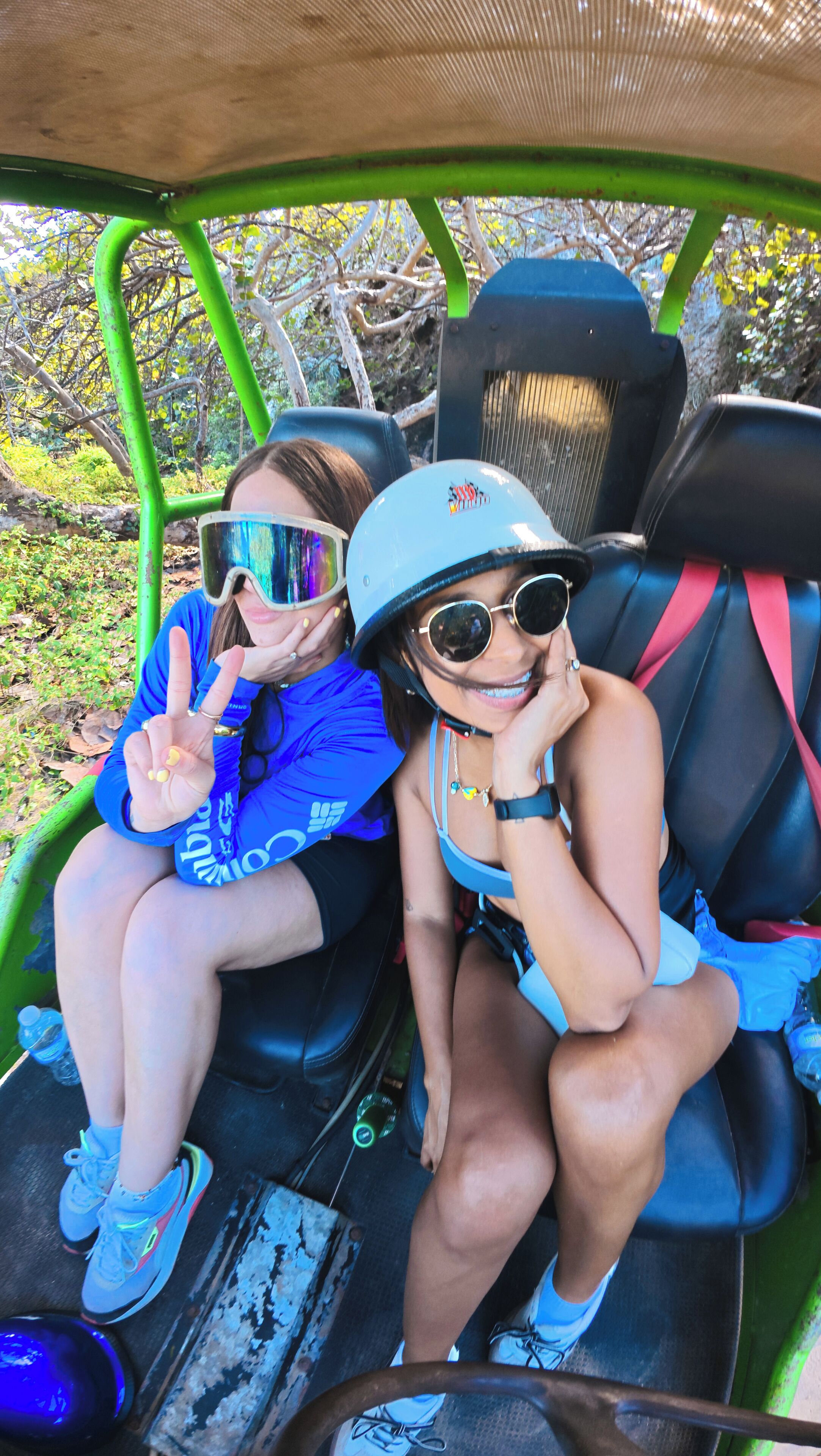 Dos personas sentadas en un vehículo de color verde, vestidas con ropa informal y gafas de sol.