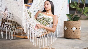 Una persona relajándose en una silla hamaca de macramé al aire libre, con suelo de concreto y una planta en maceta cerca.