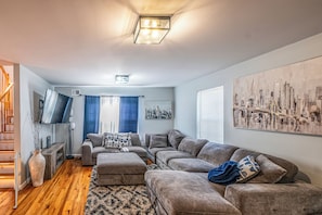 Ein Wohnzimmer mit einem Ecksofa, einem Flachbildfernseher, einem Stadtbildgemälde und einer Treppe.