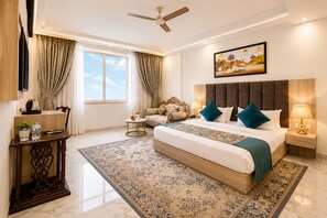 A hotel room with a large bed, a sofa, a desk with a lamp, and a painting on the wall.