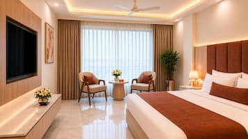 A hotel room with a bed, two armchairs, a TV, a small table with flowers, a plant, and a ceiling fan.