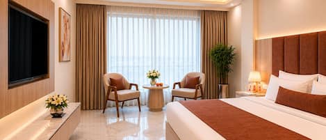 A hotel room with a bed, two armchairs, a TV, a small table with flowers, a plant, and a ceiling fan.