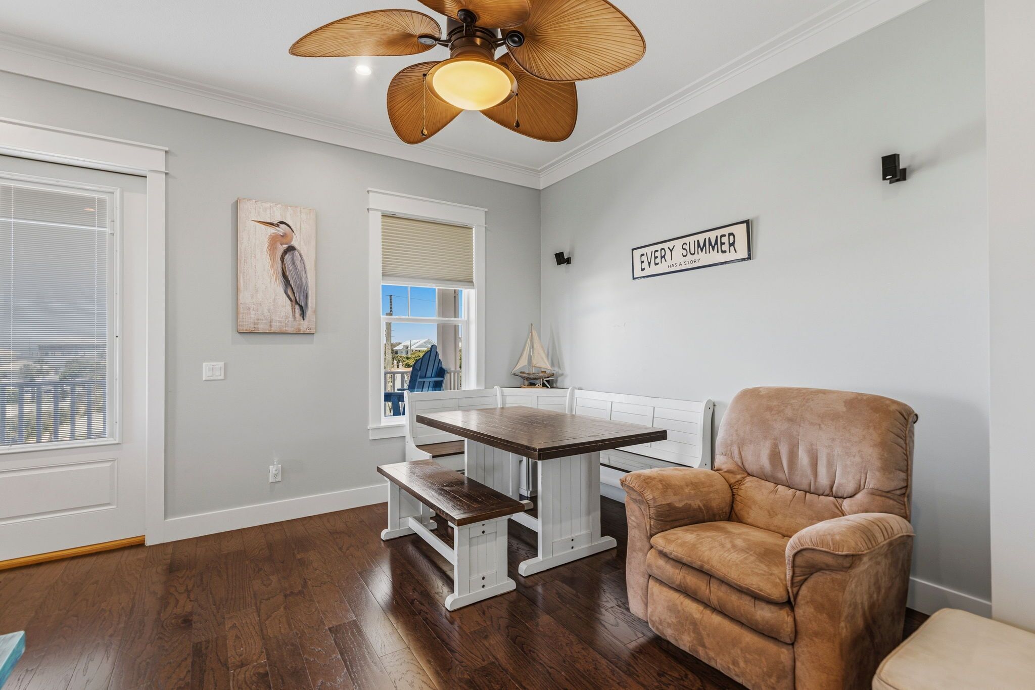 Un coin repas avec une table et un banc en bois, une chaise marron, un ventilateur de plafond et une fenêtre avec des stores.