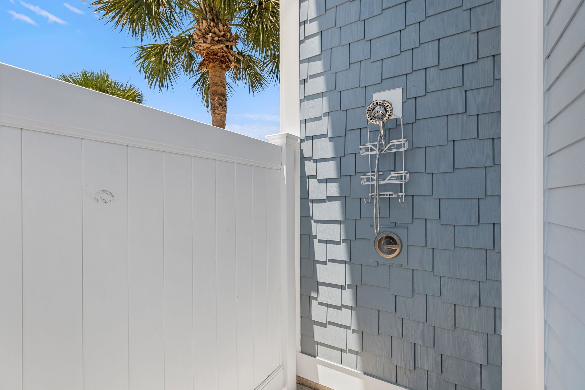 A white exterior wall with a grey shingle pattern, a metal outdoor shower, and a palm tree in the background.