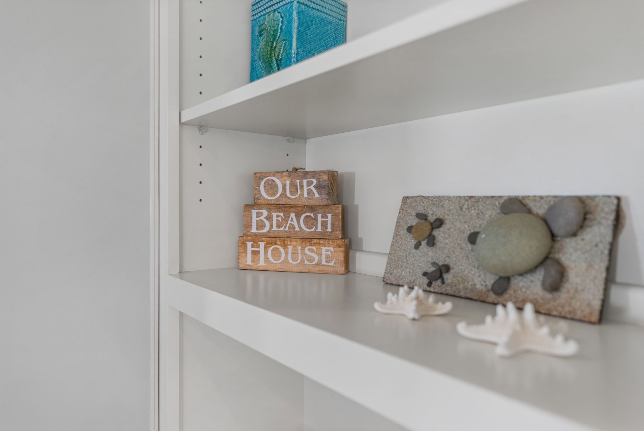 A white shelf with wooden blocks spelling 'OUR BEACH HOUSE,' a decorative rock art piece, and white coral-like decorations.