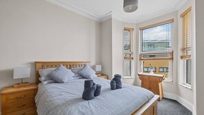 A bedroom with a bed, wooden headboard, two bedside tables with lamps, a window with blinds, and a view of a building outside. - Spacious sea view apartment, West Hoe Free Parking (Plymouth)