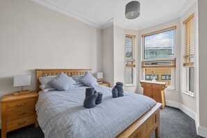 Ein Schlafzimmer mit einem Bett, einem Holzkopfteil, zwei Nachttischen mit Lampen, einem Fenster mit Jalousien und Blick auf ein Gebäude.