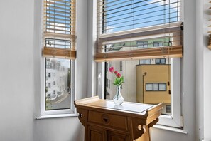 Ein Eckzimmer mit Schreibtisch aus Holz, einer Blumenvase und einem Fenster mit Jalousien.