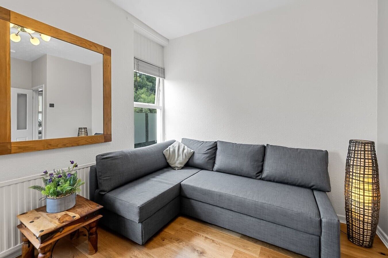 Un salon moderne avec un canapé d’angle, un miroir à cadre en bois, une petite table sur laquelle est posée une plante, et un grand lampadaire.