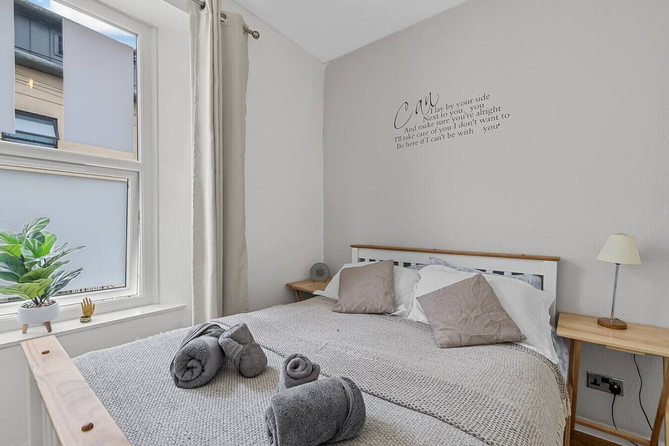 A bedroom with a bed, pillows, a bedside table, a lamp, a wall clock, a potted plant, and a window with curtains.