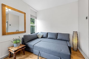 Ein Zimmer mit grauem Sofa, einem Holznachttisch mit Pflanze, einem spiegel mit Holzrahmen und einer hohen Stehlampe.