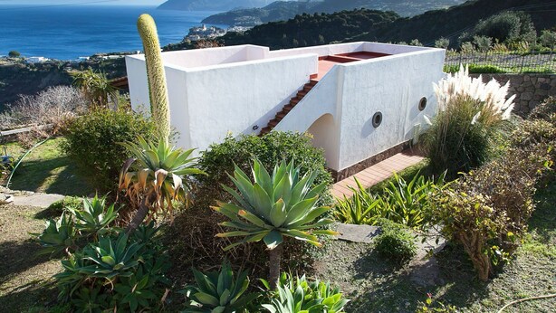 Un bâtiment blanc avec un toit rouge, entouré de différentes plantes et offrant une vue sur un paysage côtier avec des montagnes en arrière-plan.