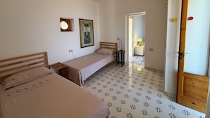 Une chambre avec deux lits simples, un ventilateur de plafond, une petite fenêtre et une porte avec un cadre en bois.