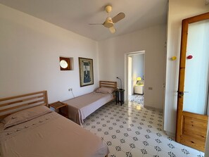 A room with two single beds, a ceiling fan, a small window, and a door with a wooden frame. - Olivetta – Vacation Home with Ocean View & Garden, Lipari (Aeolian Islands) (Lipari)
