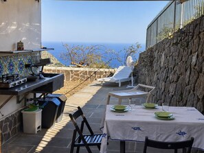 An outdoor patio with a grill, dining table, chairs, and a view of the sea. - Olivetta – Vacation Home with Ocean View & Garden, Lipari (Aeolian Islands) (Lipari)