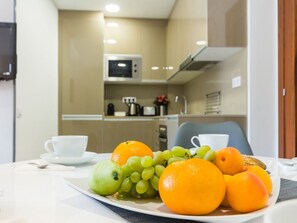 Ein Esstisch mit Früchten und einer Tasse, ein Mikrowellenherd und eine Kaffeemaschine im Hintergrund.