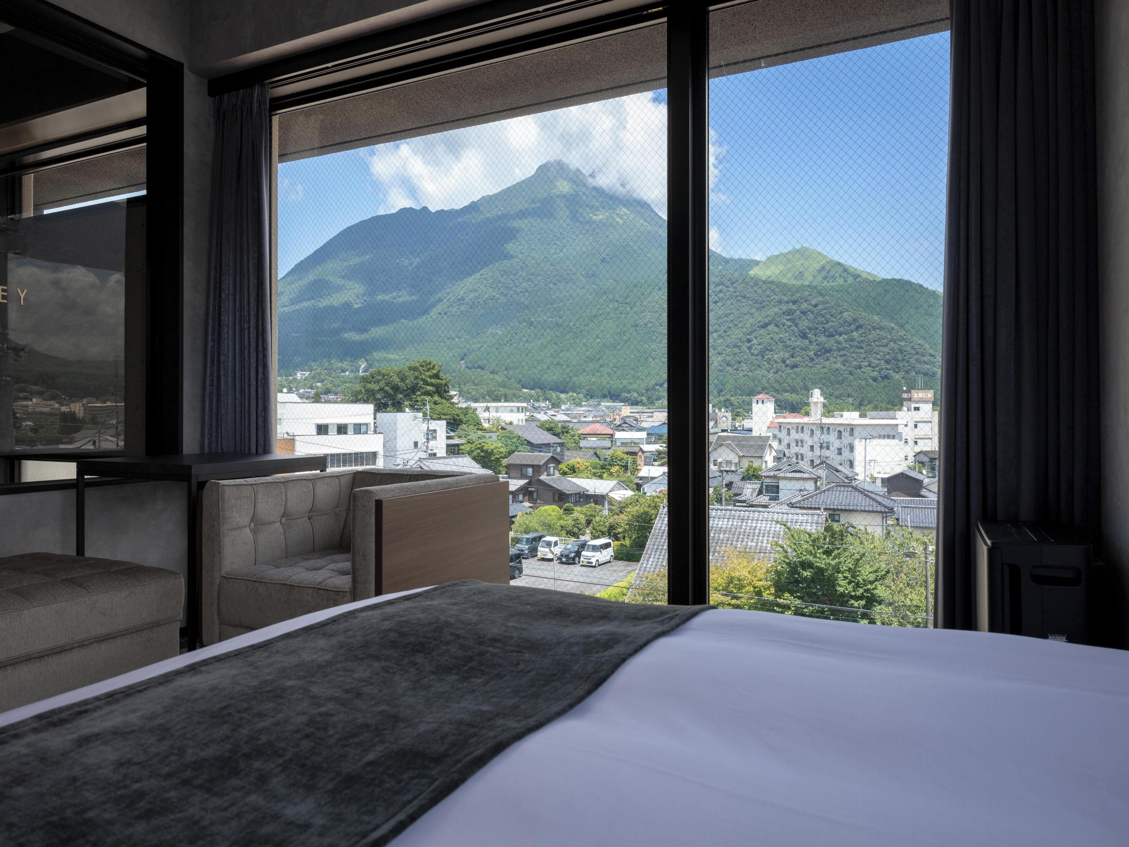 A hotel room with a large bed, a sofa, and a view of a mountain and cityscape through a large window.