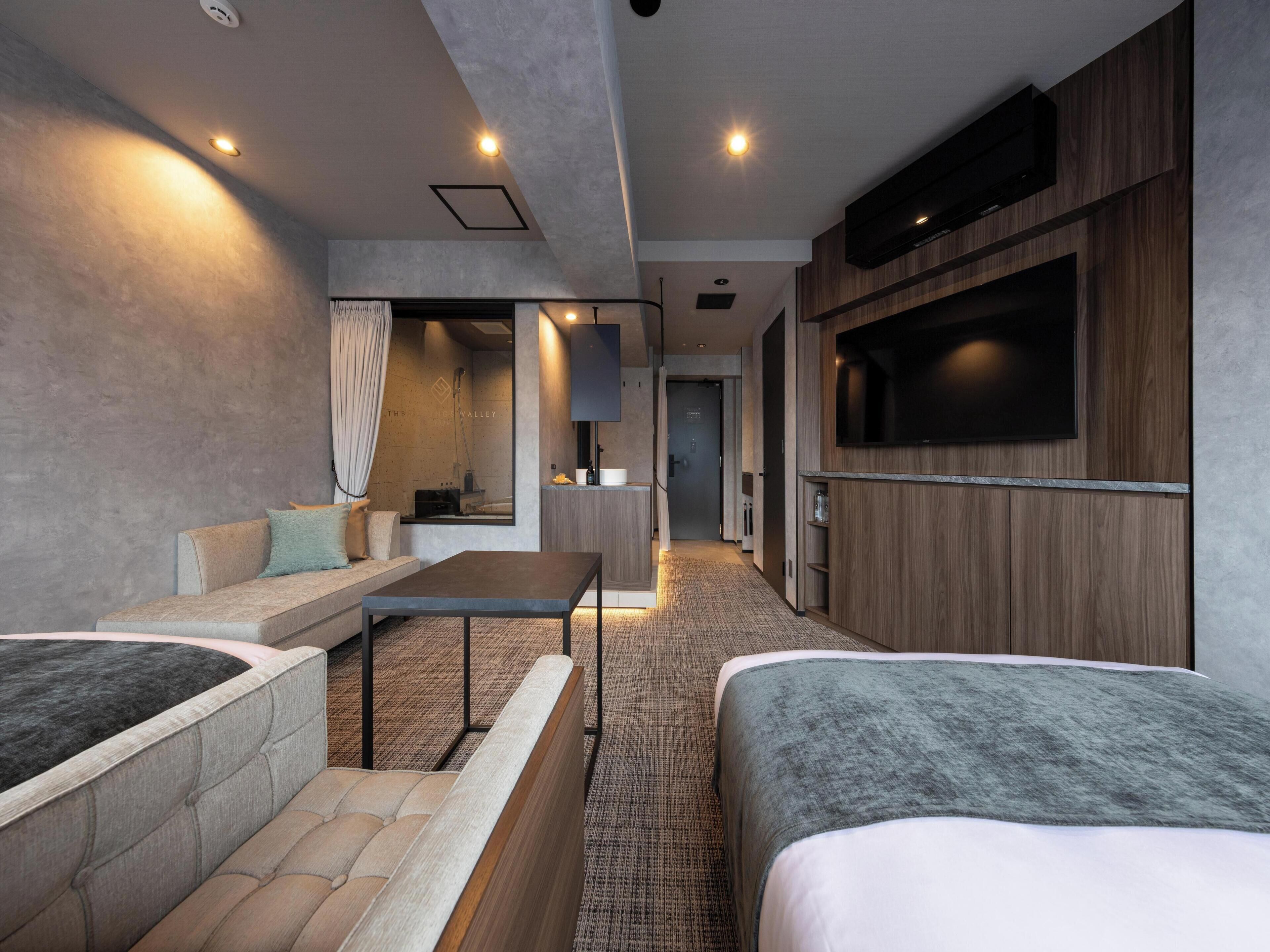 A modern hotel room with a sofa, a bed, a TV, and a bathroom visible through an open door.