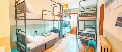 A dormitory room with bunk beds, a radiator, a window with curtains, and a small stool.