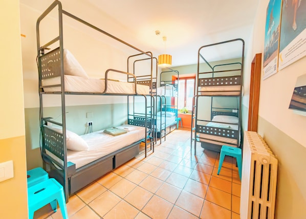 A dormitory room with bunk beds, a radiator, a window with curtains, and a small stool.
